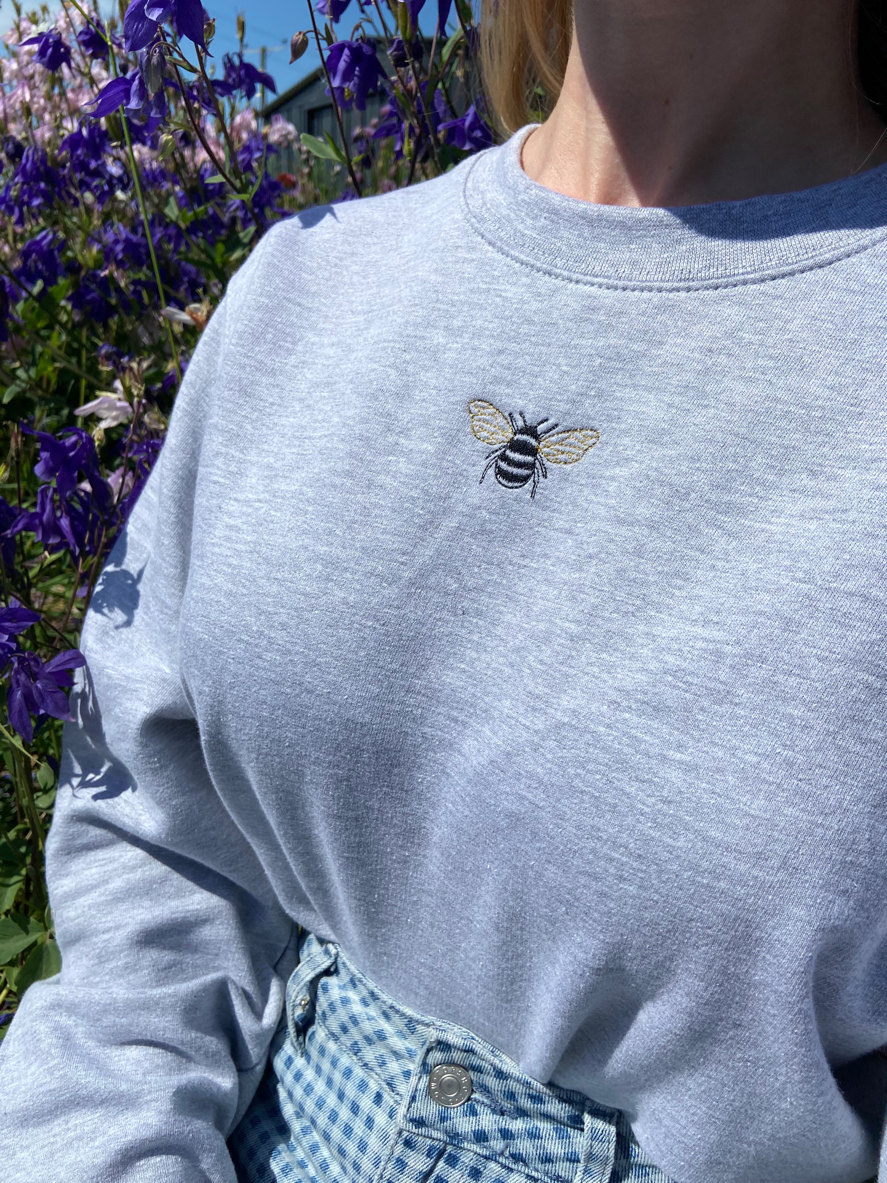 Embroidered bumble bee design on grey sweatshirt, modelled outside surrounded by wild flowers.