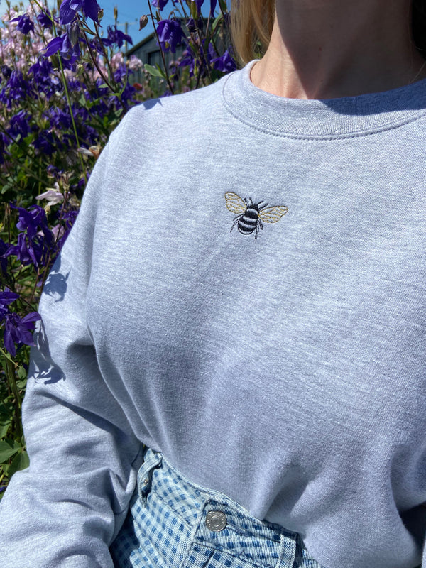 Embroidered bumble bee design on grey sweatshirt, modelled outside surrounded by wild flowers. 