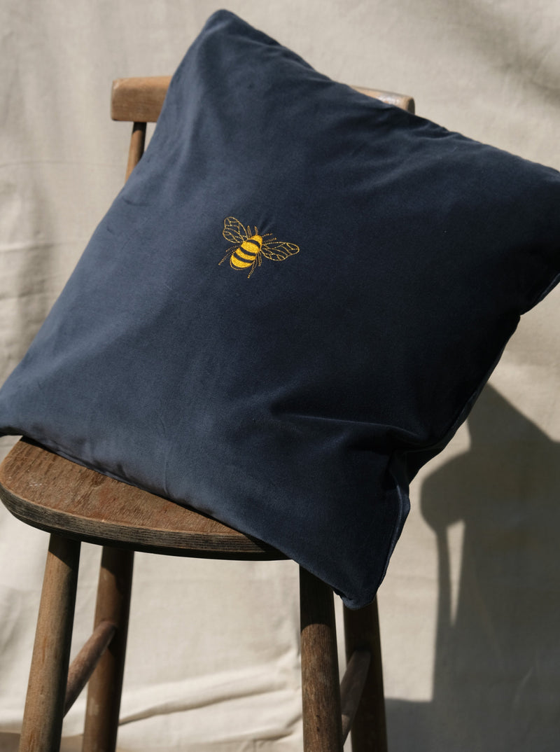 Large gold embroidered bee design onto a dark blue cotton velvet cushion cover, sitting on a wooden stall.