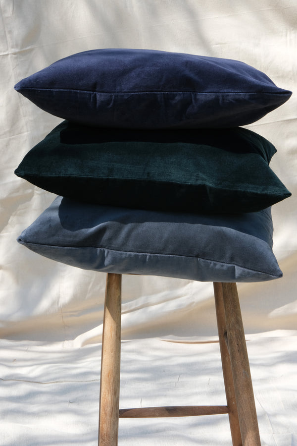 A stack of cotton velvet cushion covers, showing the various options available to buy, Dark Blue, Forest Green and Dark Grey. Sitting on a wooden stall.