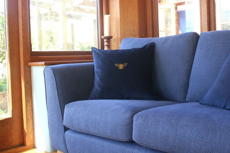 Large gold embroidered bee design onto a dark blue cotton velvet cushion cover. Sitting on a blue sofa.