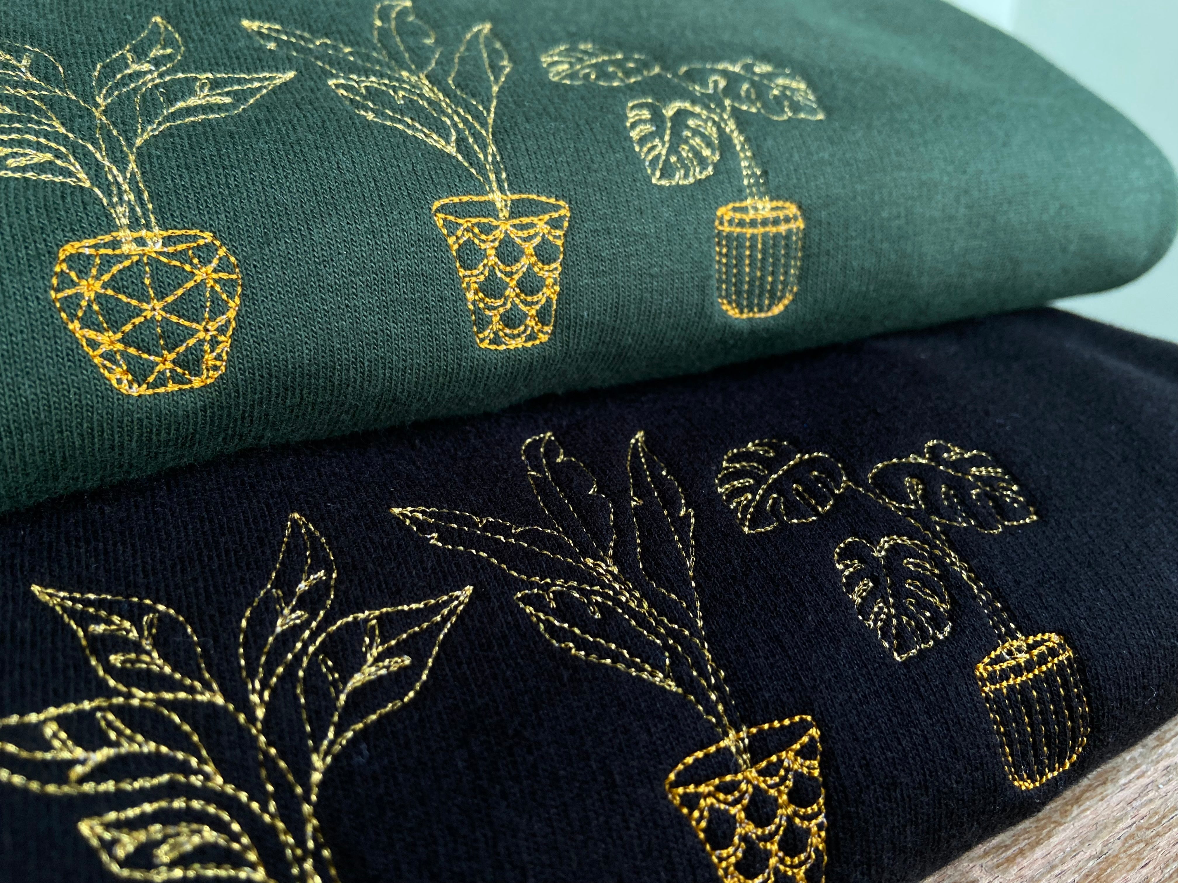 Close up of embroidered golden house plant design, stack of two sweatshirts on forest green and black.