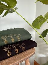 Embroidered golden house plant design, stack of two sweatshirts on forest green and black. Monstera deliciosa (Swiss cheese) plant in background.